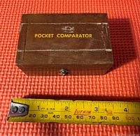 Closed wooden box with yellow text 'POCKET COMPARATOR' on top, approximately 3 inches wide shown with measuring tape below.