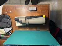 Top view of the Bushnell Spacemaster II prismatic telescope, showing the lens and body on a wooden table alongside protective case and box.