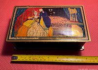 Top view showing rectangular candy tin with ornate court scene on lid and measuring tape for size reference