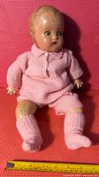 Front view of the vintage composition baby doll seated on red textured surface, showing knit pink outfit and measuring tape indicating size at about 18 inches tall.