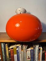 Orange rounded pendant lamp shade viewed from front, resting on shelf with books below.