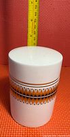 Front view of white ceramic canister with lid closed showing decorative orange and black geometric band, with a measuring tape beside illustrating about 6 inch height.