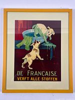 Front view of framed color print showing two dogs with green paint and text 'DE FRANÇAISE VERFT ALLE STOFFEN'. Yellow wooden frame visible.