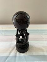 Front angle of wooden sculpture showing abstract globe with continents and supporting human figures on a round base.