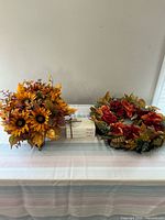 Photo showing an artificial fall floral arrangement with yellow sunflowers, orange leaves, and a fall-themed wreath with red, orange, and green leaves and flowers arranged on a table.