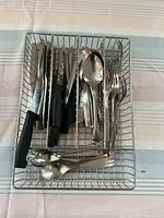 Metal wire tray containing assorted stainless steel cutlery including cutting knives, forks, tablespoons, teaspoons, and steak knives with black handles.