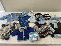 Collection of 20+ crocheted baby boy clothing items including sweaters, hats, and booties in blue and neutral shades arranged together on floor.