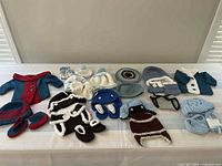Overview of multiple baby boy crocheted clothing items laid out on a bed including jackets, mittens, boots, diaper covers, and hats in colors such as blue, white, brown, and red.