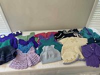 Photo showing an assortment of crocheted baby dresses and sweaters in various colors including purple, green, red, cream, lavender, and teal laid out on a table.