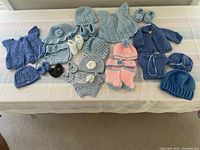 Full set of 29 crocheted baby clothing items laid out, including diaper covers, hats, booties, cardigans, and dresses, mostly in shades of blue with some peach and white accents.