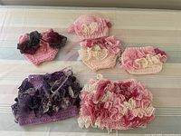 Photo showing two crocheted baby hats, multiple diaper covers, and ruffled skirts in pink, cream, and purple colors laid on a table.