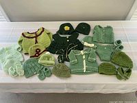 27 handmade green crocheted baby clothing items including hats, booties, sweaters and diaper covers arranged on a surface