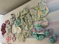 Overview of 34 baby clothing and accessory pieces laid out including sweaters, hats, booties, diaper covers, and crochet collar pieces in pastel multi-colors.
