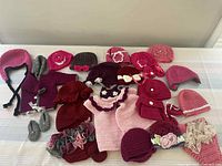 Full lot of 31 crocheted baby girl items in various shades of pink, red, burgundy, and gray including hats, diaper covers, dresses, and booties photographed spread out on a striped fabric background.