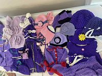 Multiple crocheted baby clothes and accessories in shades of purple and pink arranged on a flat surface showing variety of items including hats, dresses, booties, and diaper covers.