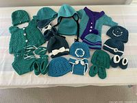 Overview of all 26 crocheted baby clothing items laid out on a flat surface, showing a variety of hats, booties, sweaters, diaper covers, mittens, and dresses in teal, blue, and purple hues.