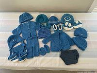 Full set of 23 crocheted baby items in shades of turquoise laid out on striped surface including hats, booties, diaper covers, pants, and cardigans.