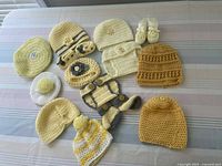 Top-down view of 11 yellow and cream crocheted baby hats, diaper covers, and booties arranged on a striped surface.