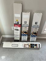 Four boxed vinyl mini blinds standing upright and lying on carpeted floor near wall outlet.
