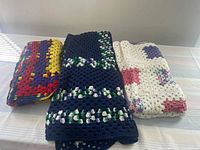 Three crocheted blankets folded and stacked. Small multicolored blanket, large navy mix blanket with green and white accents, medium granny square blanket folded at top.