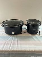 Two black slow cookers on a striped tablecloth, one large oval KitchenSmith slow cooker with glass lid, one smaller round Crock-Pot slow cooker with glass lid missing a knob.