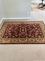 Floral oriental area rug with detailed floral and vine pattern on red center and beige border, displayed on carpeted floor.