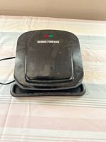 Closed black George Foreman grill on a cloth surface showing brand name and power lights on top with drip tray underneath.