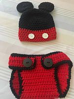 Top view of black and red crocheted baby beanie hat with mouse ears and white button accents, alongside matching red diaper cover with black trim and black buttons.