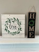 Two wall signs on a table against a gray wall: a white square sign with a green leafy wreath and 'love is all you need' text, and a tall black rectangular HOME sign with a wreath in place of the O and hanging rope.