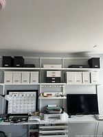Wide view showing 11 medium white and 5 black heavy-duty cardboard organizer boxes on shelves above desks and monitors.