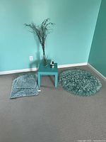 Overall view of corner showing small light blue table with items on top, round shaggy rug to side, two textured cushions on floor, and glass vase with branch on table.