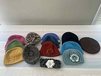 All 13 crocheted hats and 1 crocheted headband arranged on a table, showing variety of colors and presence of flower decorations on two hats and the headband.
