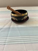 Side view of black metal Tibetan singing bowl with gold and copper accents, containing wooden mallet, sitting on round cushion.