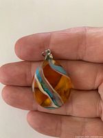 Front view of teardrop glass pendant showing amber body with blue and cream ribbons and metal bail