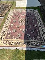 Top-down photo of a rectangular area rug with a floral medallion in the center, primarily pink with cream and black borders and floral accents, laid on grass with natural light and some shadow obscuring part of the rug.