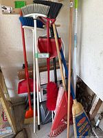 Overall assortment of cleaning and gardening tools including brooms, shovels, rake, and step ladder leaning against the wall.