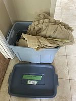 Blue plastic tote filled with beige curtains and gold curtain rods, placed on tile floor near door.