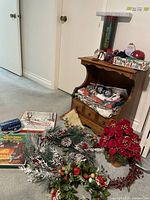 Photo of the side table with folded Christmas tablecloths or fabric on its lower shelf, silver Christmas ornaments inside the drawer, pinecone and berry Christmas wreath on the floor, Christmas records, a Christmas stocking, and boxes arranged on the floor beside it.