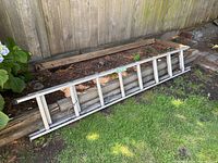 Full length side view of aluminum ladder resting horizontally on grass and wood logs outdoors, showing entire ladder length.
