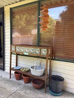 Full view of the elevated wooden planter box with floral tiles, three terracotta pots, one large black plastic pot, blue decorative plate, white wicker basket on wire support, and wooden wind ornament hanging above.