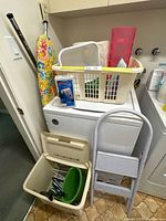 Photo showing laundry hamper, basket with various laundry items, ironing board with floral cover, and step ladder leaning on washer.