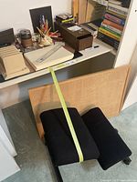Photo showing black kneeler chair, corkboard, wooden file/card holder, ink stamp, pencils, pen holders, and stacked books on shelves.
