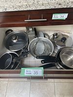 Drawer with assorted pots, pans, strainers, metal box grater, and multiple lids showing variety of cookware sizes and types.