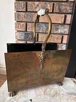 Front view of brass magazine rack showing curved brass handle with ceramic grip and lion paw feet.