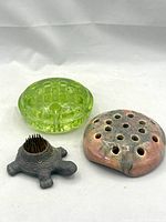 Three vintage flower frogs arranged together: bright green uranium glass piece, pink/beige glazed pottery frog with multiple holes, and dark cast lead turtle-shaped frog with metal pins.