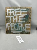 Front view of street art stencil painting on recycled cardboard featuring 'FREE THE PEOPLE' in silver paint over patterned brown cardboard.