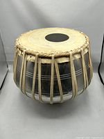 Front and side view of Indian bayan drum showing metal body with decorative stripes, natural hide tension straps, and goatskin drumhead with black syahi patch.