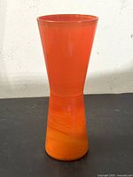 Photo of an orange hourglass slag glass vase on a dark surface against a white wall, showing the smooth and glossy texture with subtle swirl patterns.
