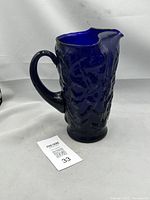 Front side view of cobalt blue glass pitcher showing textured surface and integrated handle on a plain background