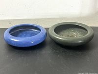 Pair of vintage low-form pottery bowls on black surface. Left bowl is vibrant blue with glossy finish. Right bowl is matte green. Both have broad rims and shallow profiles.